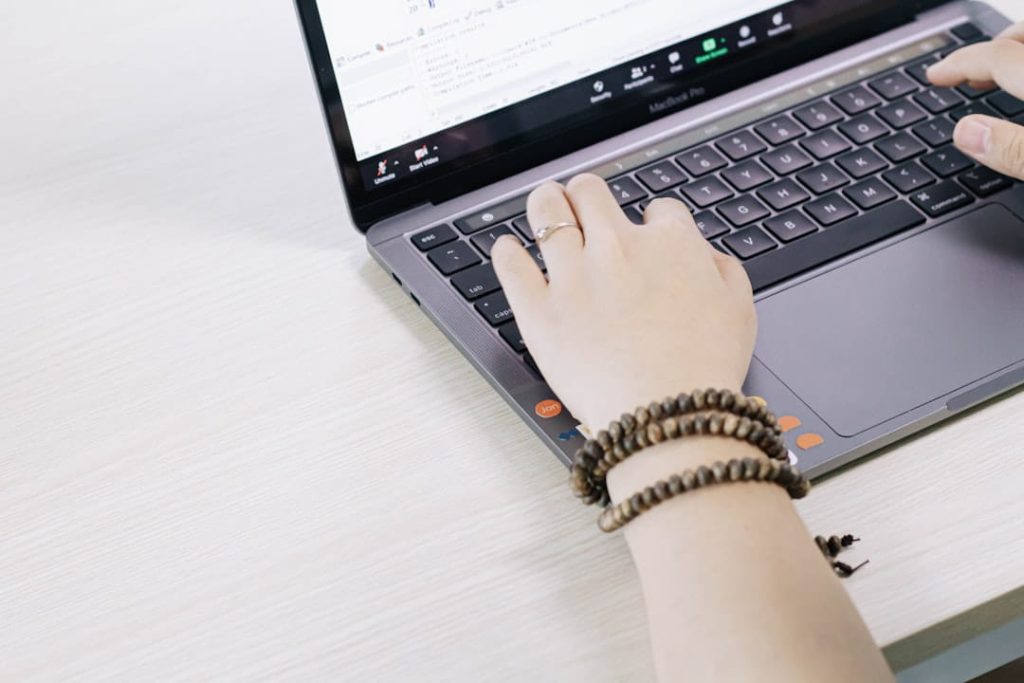 A close-up of hands typing on a silver MacBook Pro laptop on a light wooden desk. The screen displays a programming interface or video call window. The left hand wears a beaded bracelet, and the laptop features colorful stickers, including one with "Jon." The scene suggests a work or coding environment, perfect for researching how to follow up after a networking event.