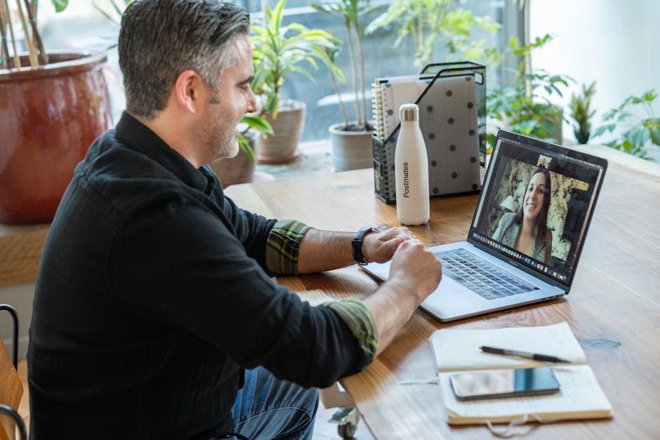 A man on a video call with a woman, discussing how to start a business while seated at a wooden table with a laptop, notebook, and a branded water bottle in a plant-filled room.