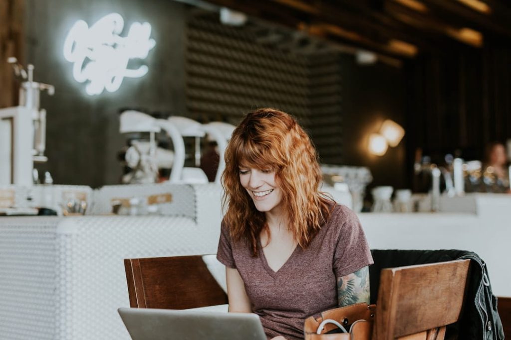 A smiling red-haired woman works on her laptop in a cozy coffee shop, capturing the flexible and independent spirit of how to start a small business on your own terms.