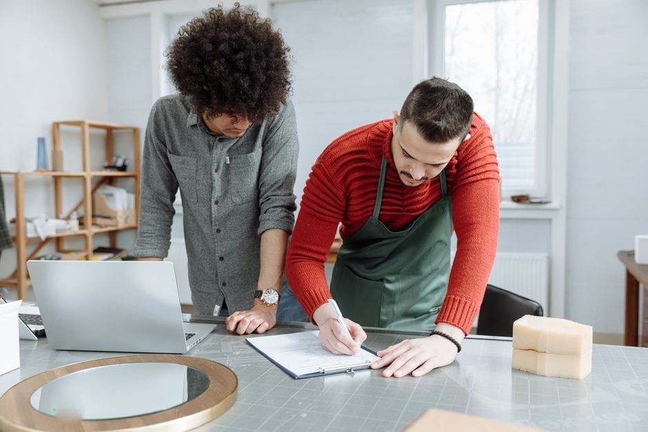 Two entrepreneurs review a clipboard and laptop in a bright craft workshop, reflecting the hands-on planning that goes into how to start a small business from the ground up.