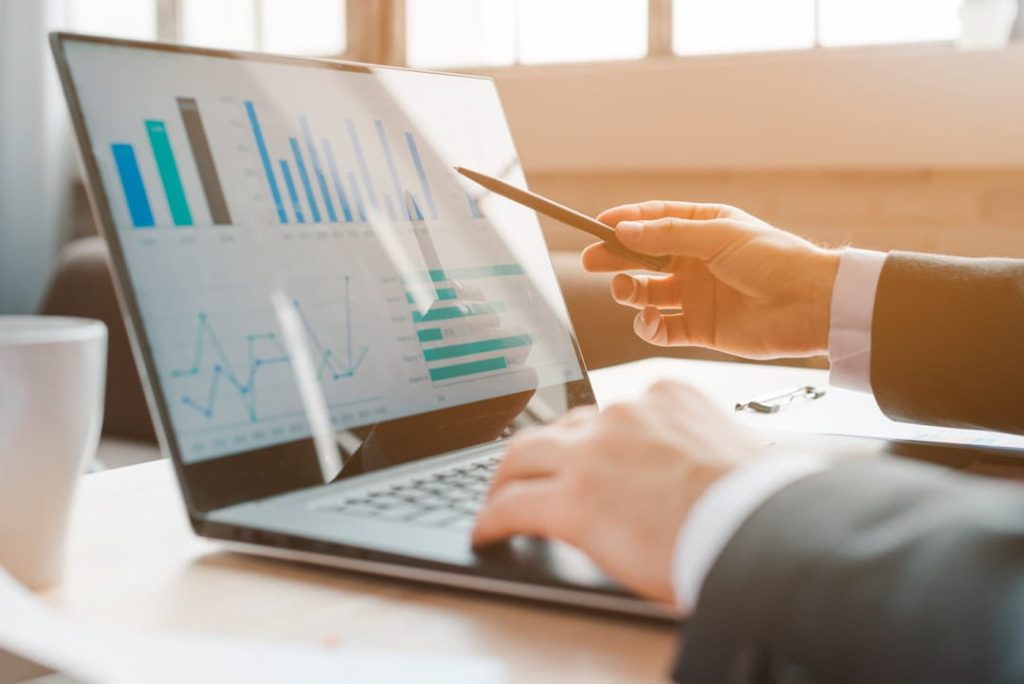 A suited professional points at a multi-chart analytics dashboard on a laptop in a sunlit office, highlighting the data-driven decisions behind how to start a small business successfully. 