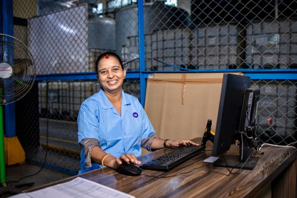 A smiling woman in a blue uniform manages inventory at a computer station inside an industrial warehouse, showing one of the many operational realities of how to start a small business.