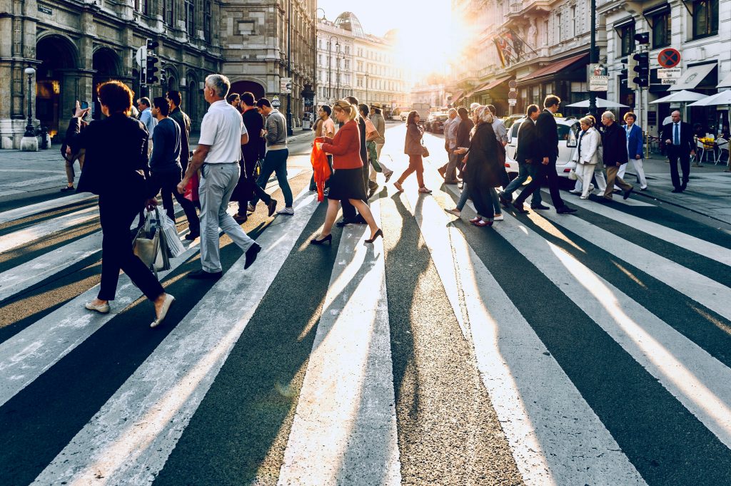 An urban street scene in Vienna during late afternoon or early evening, featuring a diverse group of pedestrians crossing a wide, striped crosswalk. The low sun casts long, dramatic shadows across the street, highlighting the bustling activity. Ornate historical buildings line the left side, while modern buildings with outdoor cafes and flags are on the right. A 'HOTEL' sign and 'Opernpassage' signage are visible, along with a few vehicles in the background. The warm sunlight creates a vibrant and dynamic atmosphere, reminiscent of the energy found at networking events Orlando.