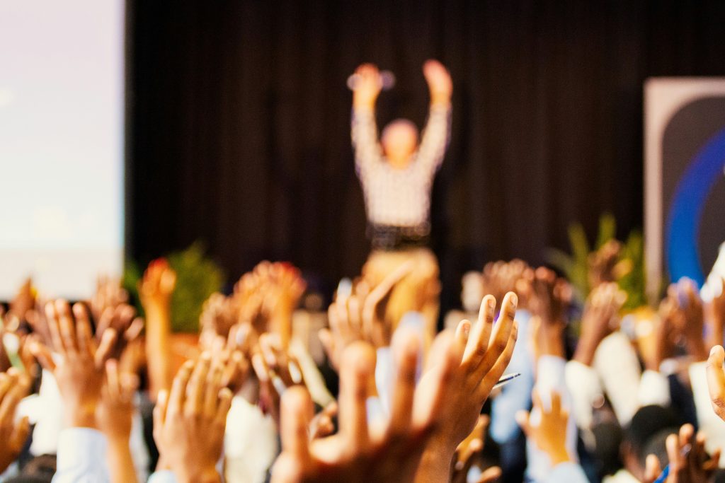 A group of diverse individuals enthusiastically raise their hands in the foreground, symbolizing active participation. In the background, a blurred figure with raised arms appears to be addressing the crowd, set against a dark curtain and hints of greenery. The setting suggests a formal event, such as a conference, seminar, or networking events San Diego, emphasizing engagement and unity.