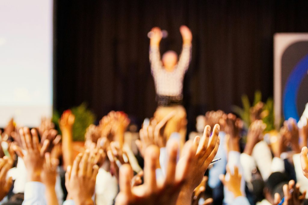 A dynamic scene showing numerous raised hands in the foreground, suggesting an engaged audience participating in an event. In the background, a slightly blurred individual with arms raised appears to be addressing the crowd. A partially visible screen on the left hints at a presentation or lecture, while warm lighting highlights the interactive atmosphere. The darker background and stage-like setup suggest a formal or educational event, such as networking events Houston, a seminar, or a conference.