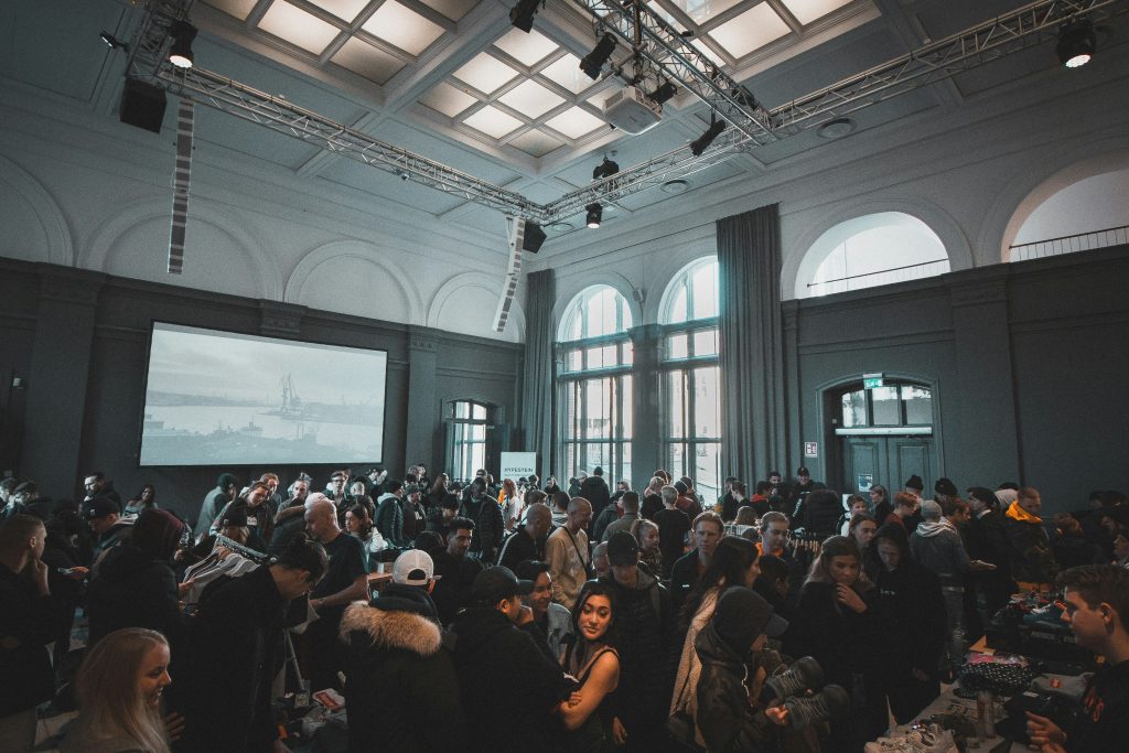 A large indoor event space with high ceilings, featuring a crowd of people mingling and browsing items. The venue has a grid-patterned ceiling with suspended truss systems holding spotlights, speakers, and a projector. A large projection screen on the left displays a monochrome industrial waterfront scene. On the right, tall arched windows with dark curtains let in natural light, and a mezzanine with metal railings is visible above. The crowd, dressed casually in hoodies, jackets, and hats, suggests a lively and informal gathering, possibly a market, convention, or one of the networking events in DC. The overall atmosphere is bright and bustling, with muted tones of gray, blue, and black.