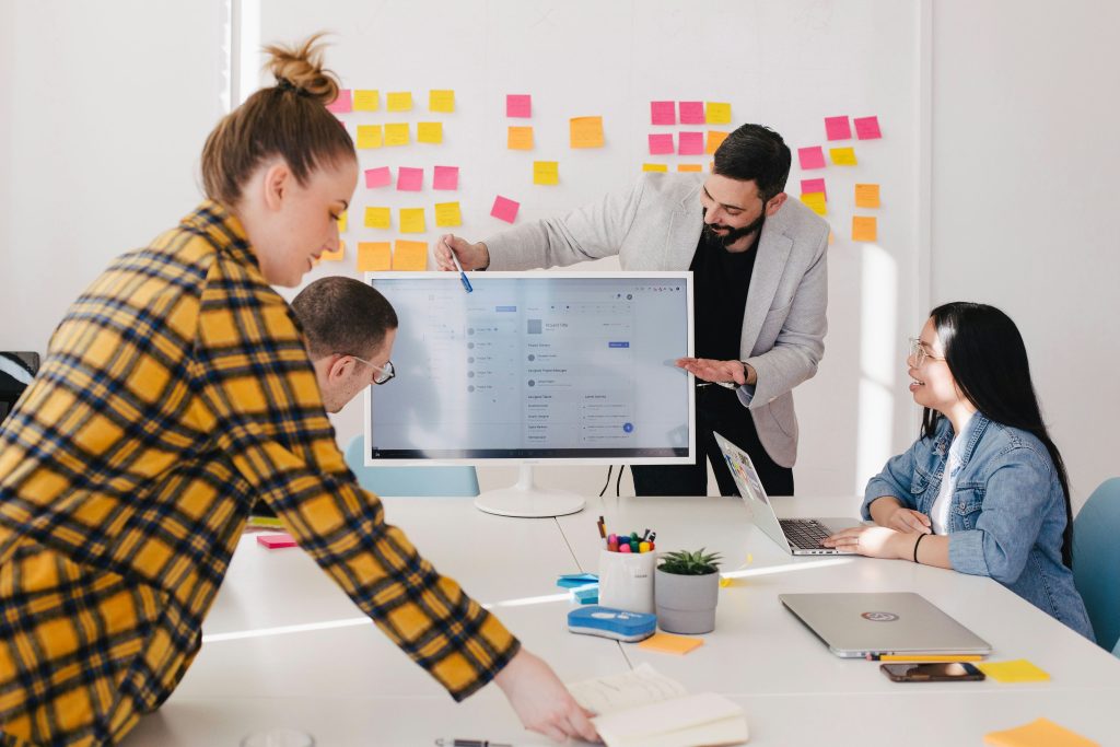 Four individuals are collaborating in a modern office setting around a white table. A central monitor displays a project management interface, while a person in a yellow plaid shirt leans over the table. Another individual in a light gray jacket stands and points at the screen, while a person in glasses observes. A fourth person, seated on the right in a denim jacket, works on a laptop. The table holds various items, including a mug with pens, a small plant, sticky notes, and a smartphone. Brightly colored sticky notes are arranged on the wall in the background, suggesting brainstorming or planning activities. The scene conveys a dynamic and creative small business consulting team environment.