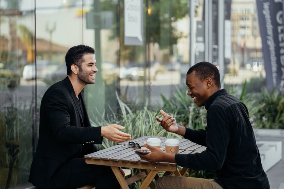 Two men enjoying a cheerful conversation at an outdoor cafe table while discussing how to build a job description template.