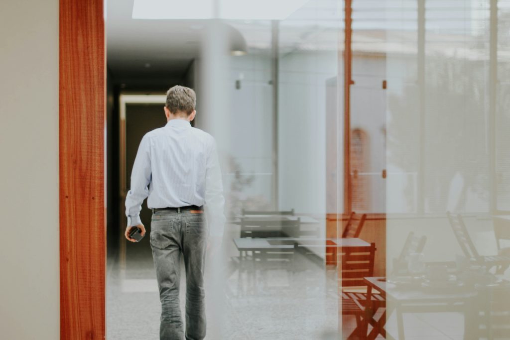 A man in a light blue shirt and grey trousers walking down a modern indoor corridor with reflective glass surfaces and visible furniture, suggesting an office or professional setting, possibly heading to discuss exit interview questions.