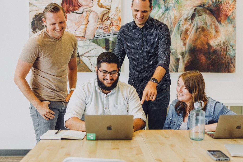 Four people collaborate around a wooden table in a bright office space. A seated man in a light button-down shirt focuses on a silver laptop, while a standing man in a dark polka-dot shirt leans over, pointing at the screen. A woman with reddish-brown hair, seated to the right, smiles while looking at the laptop, and a man in a beige t-shirt stands on the left, smiling with one hand in his pocket. The table holds a notebook, a water bottle, and a smartphone. Abstract paintings with warm and cool tones decorate the white wall in the background, creating a positive and collaborative atmosphere for an employee benefits corporation.