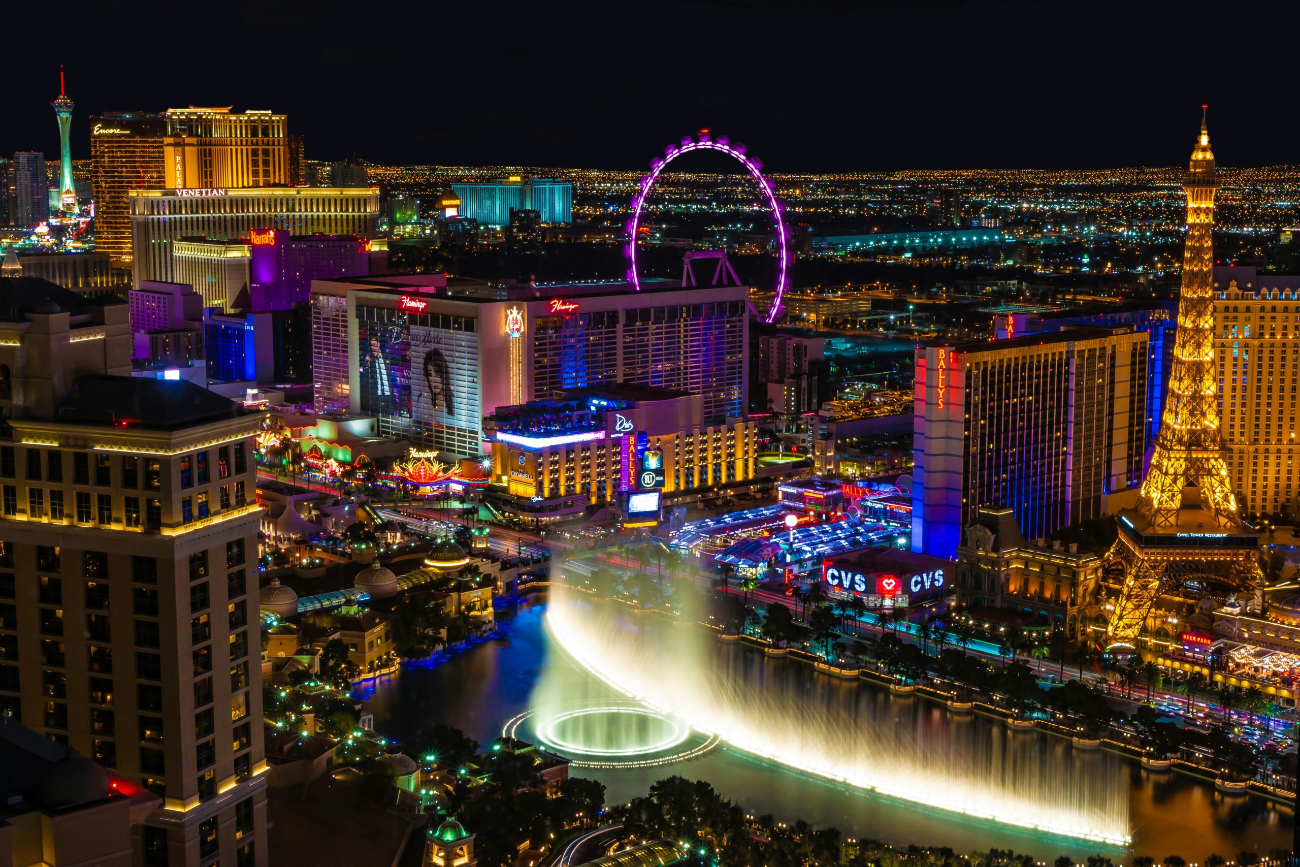 A vibrant nighttime view of the Las Vegas Strip featuring iconic hotels like The Venetian, Harrah's, Flamingo, and Bally's. A brightly lit fountain display is in the foreground, with neon lights in purple, red, blue, and yellow illuminating the scene. A Ferris wheel glows in purple in the background, and a replica of the Eiffel Tower stands prominently on the right. The bustling atmosphere highlights the city's entertainment, tourism appeal, and networking events Las Vegas.