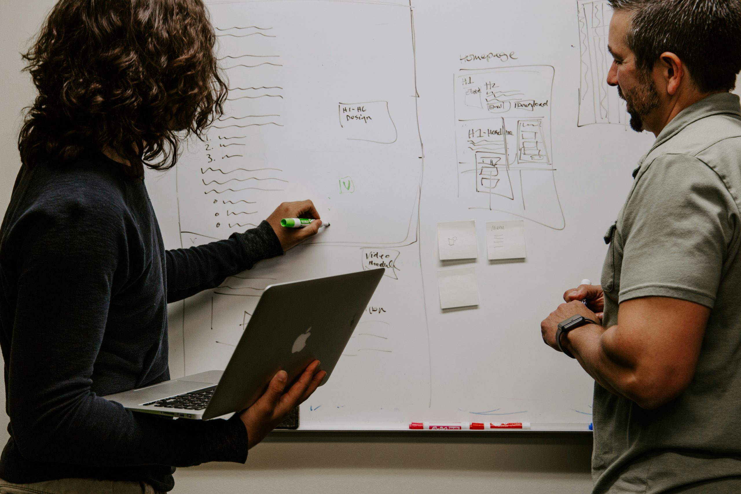 Two individuals collaborate at a large whiteboard in a professional setting. The whiteboard is filled with handwritten notes, lists, and diagrams, including a prominent wireframe labeled 'Homepage' with sections like 'H1', 'H2', 'Email', and 'Download'. One person, on the left, holds a green marker and a silver MacBook while writing on the board. The other person, on the right, observes, wearing a light olive-green shirt and a smartwatch. Sticky notes with sketches, including a cup and a menu icon, are affixed to the board. The scene captures a brainstorming session focused on website or user interface design, guided by a Client Success Manager.