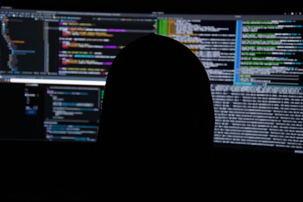 The image shows a dark, atmospheric scene featuring the silhouette of a person seated in front of a glowing computer screen, deeply engaged in what could be information management for small business. The silhouette, positioned in the lower-right corner, is entirely black, obscuring any facial details. The computer screen displays blurred lines of colorful code in shades of white, green, blue, purple, and orange on a dark background, with sections resembling a code editor or terminal. The screen is the primary light source, casting a soft glow in the otherwise dark environment. The overall composition emphasizes the act of coding or working with data, evoking themes of technology, focus, and solitude.