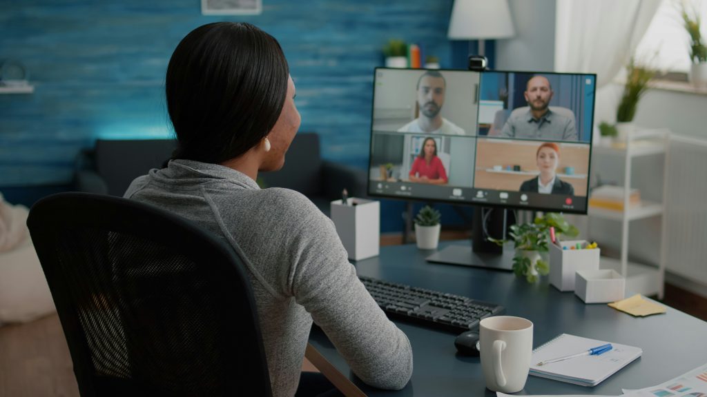 A woman is in a virtual meeting etiquette video conference call in her home office. On her desktop monitor, four participants are visible in a grid. The organized desk holds a keyboard, mouse, mug, and notebook. The room, lit by natural light, contains a blue wall, a sofa, shelving, a lamp, and several plants.