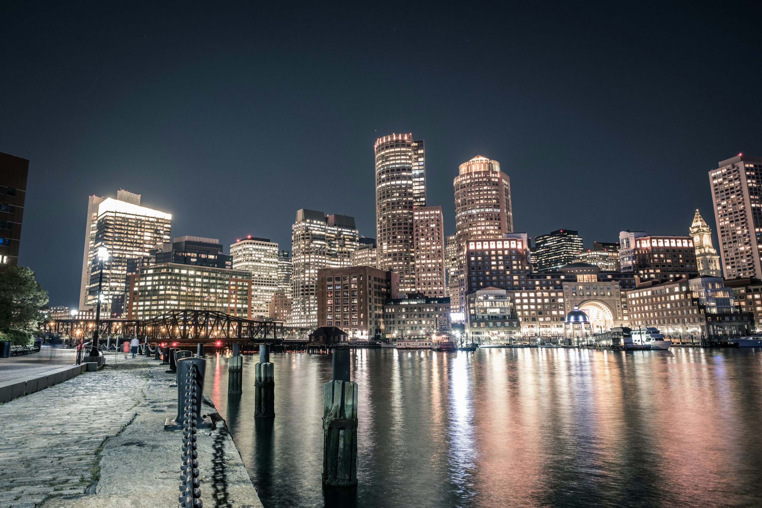 A panoramic night view of a city skyline reflected in calm waterfront waters. The skyline features a mix of modern skyscrapers and traditional architecture, illuminated with warm yellow and white lights, perhaps after a day of networking events Boston. A paved waterfront path with bollards and faint figures of people runs along the water's edge, while a metal truss bridge and docked boats add depth to the scene. The dark night sky contrasts with the vibrant city lights and their shimmering reflections on the water.