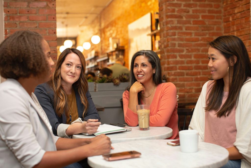 Four women sit around two small, round white tables in a cozy cafe with exposed brick walls and warm lighting. They are engaged in conversation, smiling and interacting, perhaps discussing what are angel matchmaking services. One woman holds a pen and has a notebook open, another has a tall glass of iced coffee, and a third has a white mug and a smartphone on the table. The background features shelves with decor, circular light fixtures, and a blurred figure in the distance, suggesting a casual and collaborative atmosphere.
