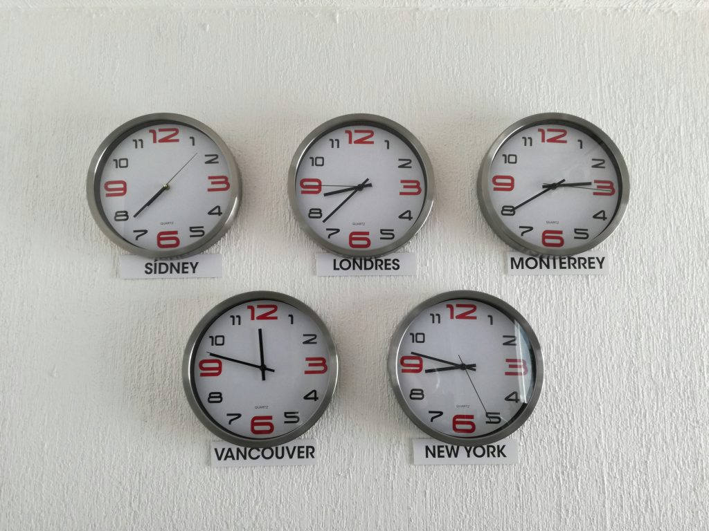 Five analog wall clocks on a textured white wall show the time in Sydney, London, Monterrey, Vancouver, and New York. The clocks feature silver frames, white faces, and red numbers at 12, 3, 6, and 9, with the other numbers in black. Each clock is labeled with its city name and arranged in a triangular pattern, creating a professional display of global time zones, highlighting the importance of virtual meeting etiquette across different regions.