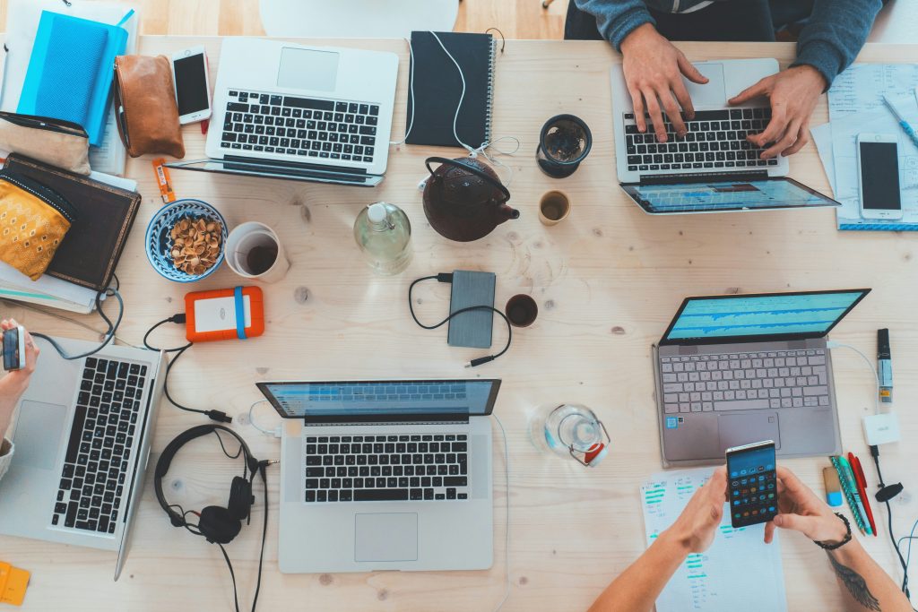 Top-down view of a light wooden table with multiple laptops, smartphones, handwritten notes, and snacks, suggesting a collaborative workspace. Five laptops, including silver MacBooks and a pink ASUS laptop, are surrounded by items like a cast-iron teapot, a bowl of pistachios, notebooks, and chargers. Hands are visible typing, holding smartphones, or interacting with devices. The scene conveys a busy, shared work or study environment with a mix of technology and personal touches like drinks and snacks, perfect for discussing the best time to post on social media.