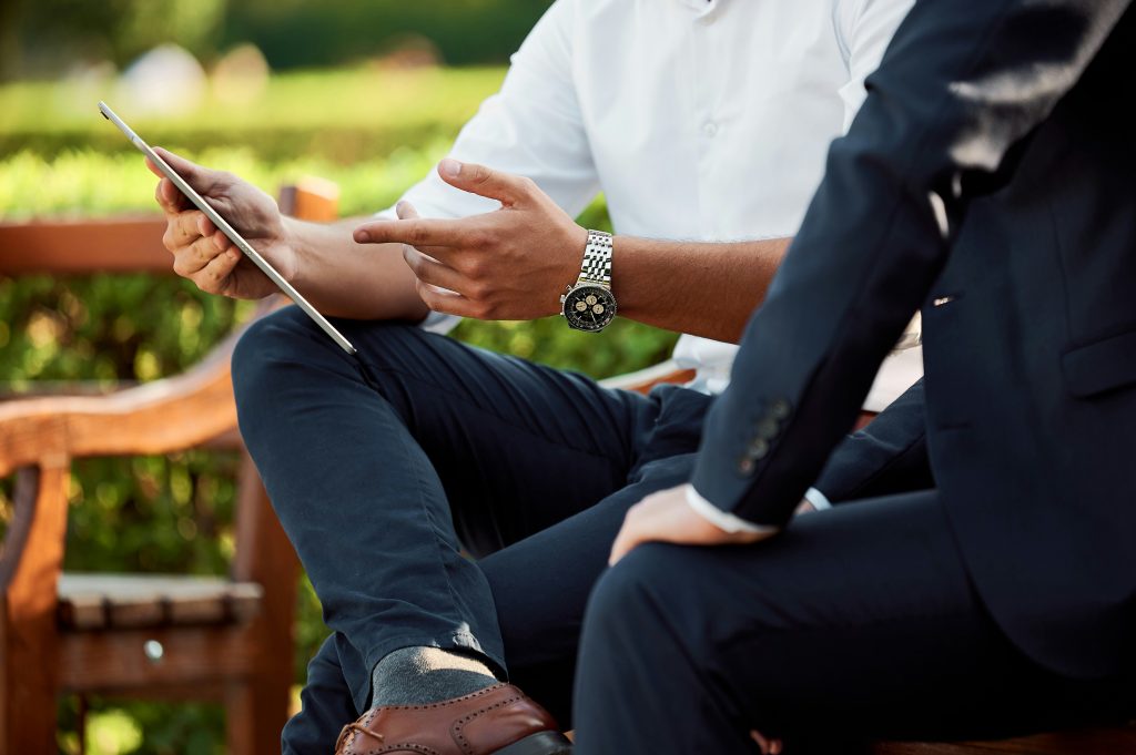 A close-up view of two men seated outdoors, focusing on their hands and lower bodies. The man on the left, wearing a white shirt and dark blue trousers, holds a silver tablet in one hand and gestures towards it with the other. He wears a silver wristwatch and brown leather brogue shoes. The man on the right, dressed in a dark blue suit, has his hand resting on his thigh. The blurred background features green foliage and wooden structures, suggesting a park or garden setting. The scene subtly conveys what are angel matchmaking services, as part of a professional yet casual interaction.