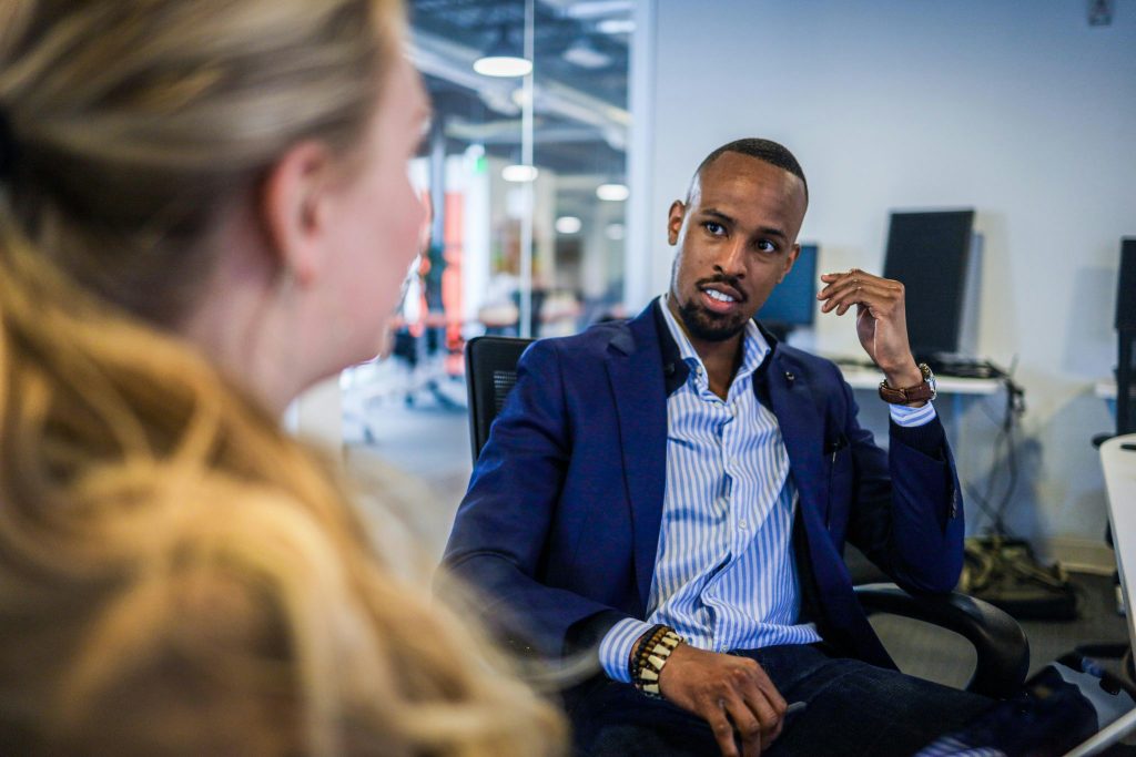 Three individuals are seated around a large wooden table in a bright, plant-filled room, perhaps discussing how to find angel investor Los Angeles. A woman on the left, wearing a beige hoodie, has a black laptop in front of her and appears engaged in the conversation. A man in the center, with dark skin, a beard, and a black t-shirt, gestures while holding a light blue mug. Another man, seen from behind, listens attentively, wearing a reddish-brown t-shirt and holding a similar mug. The background features a large bay window with white frames, potted plants, and outdoor greenery visible through the glass, creating a casual and collaborative atmosphere.