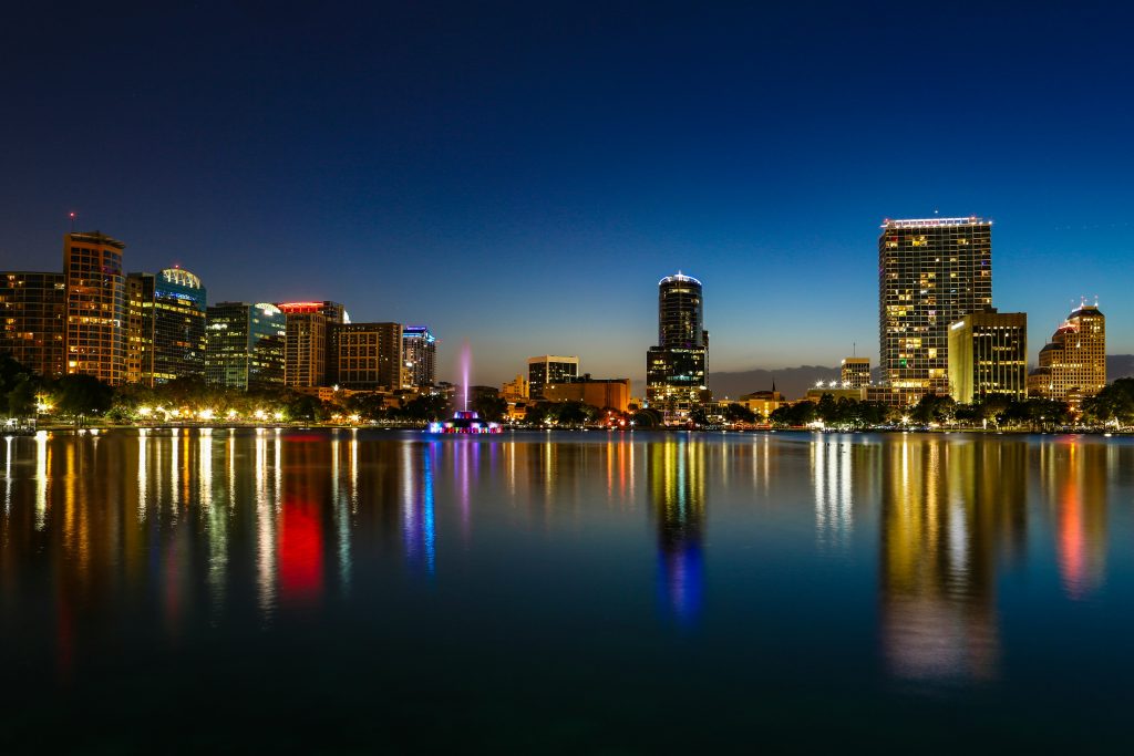 A vibrant nighttime city skyline reflected in a calm body of water. The scene features illuminated buildings with warm yellow, orange, blue, and red lights, a colorful fountain in the center, and shimmering reflections on the water. The dark blue sky contrasts with the bright city lights, creating a tranquil and picturesque urban landscape—perfect for networking events Orlando.