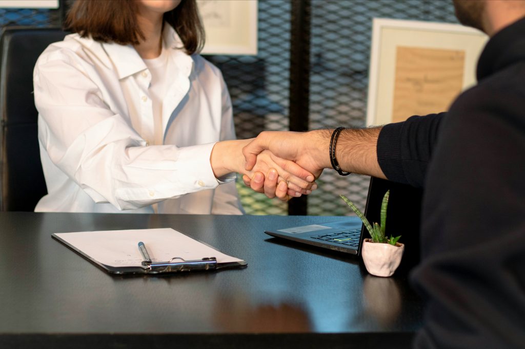 Close-up of a professional handshake between two individuals across a dark wooden table in a modern office setting. The person on the left wears a white button-up shirt, while the person on the right wears a dark sweater and a braided bracelet. The table features a clipboard with a pen, an open laptop, and a small potted succulent plant. The background includes a textured wall and framed documents, adding to the professional atmosphere, similar to networking events Miami.