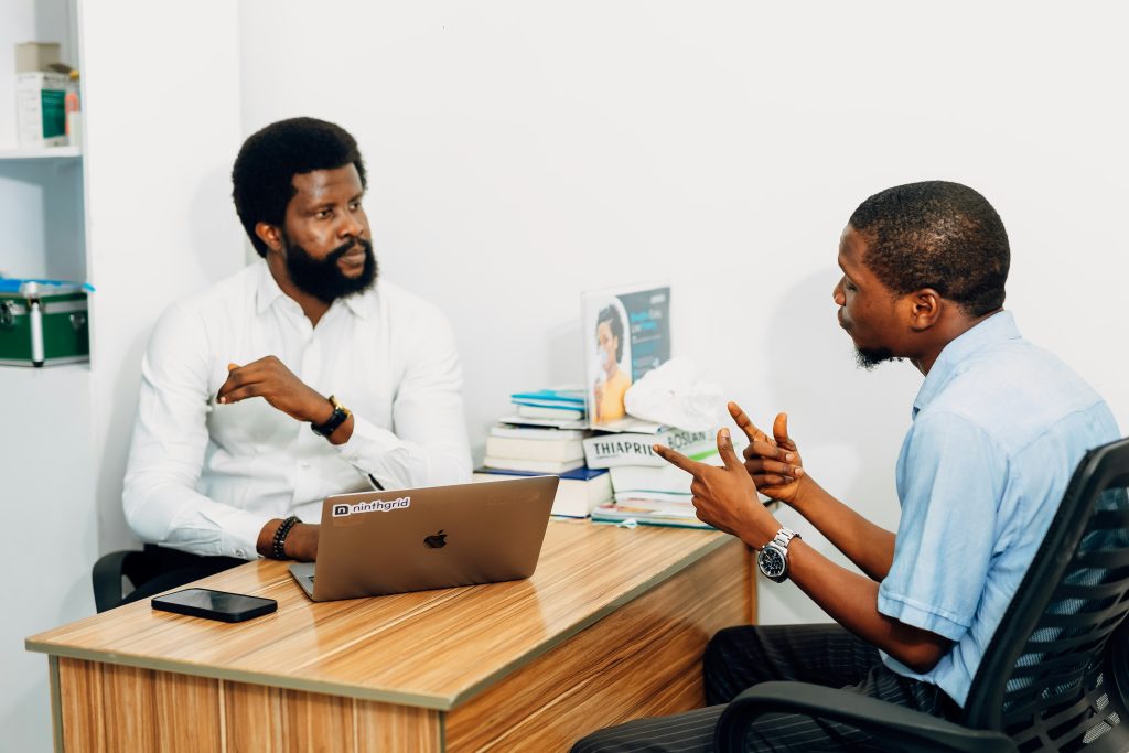 Two individuals seated at a wooden desk in an office setting, one in a white shirt listening and the other in a light blue shirt gesturing while speaking, with a laptop, smartphone, exit interview questions, and books on the desk.