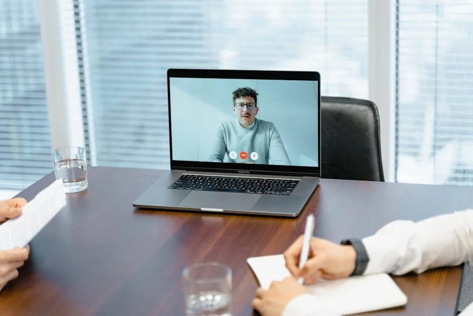 A MacBook Pro on a dark wooden table displaying a video call with a man wearing glasses and a light-colored top. Two in-person participants are partially visible, one holding papers and the other writing in a notebook. The setting is a bright office with a large window and blinds in the background, perfect for online business networking events.