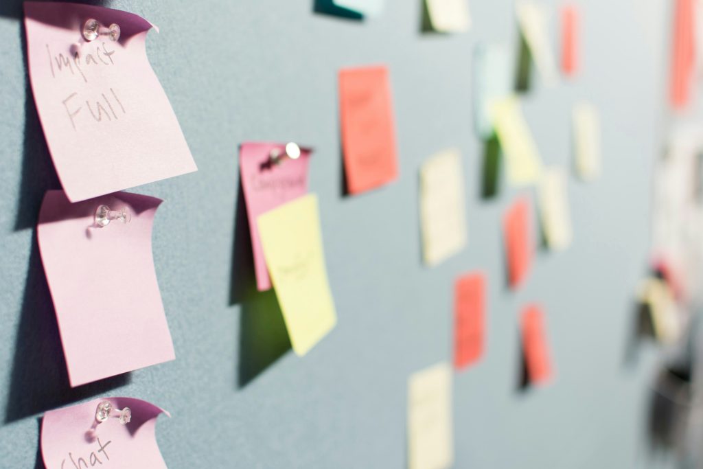 Close-up of a bulletin board with pink sticky notes in focus on the left. The top sticky note reads 'Impact Full,' the middle is blank, and the bottom partially shows the word 'chat.' The notes are pinned with clear thumbtacks. The background features blurred multicolored sticky notes, suggesting a brainstorming or collaborative workspace created by a small business consultant.