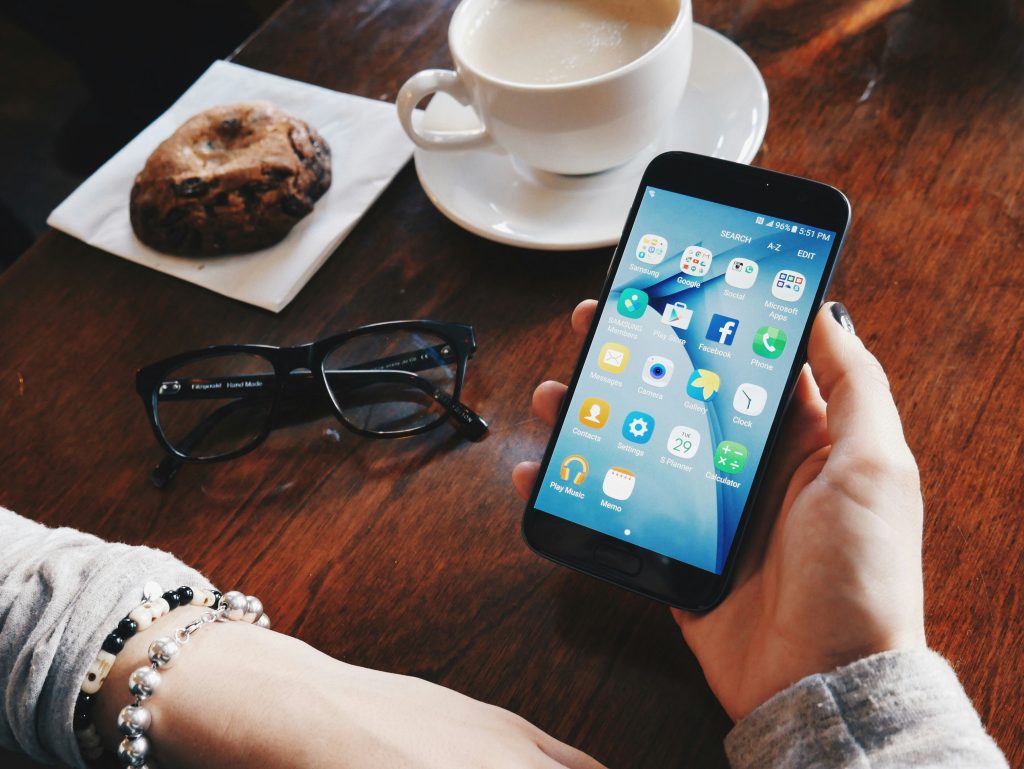 Overhead view of a dark wooden table featuring a black smartphone held in a hand with dark nail polish, displaying an organized Android home screen with app icons and folders. Surrounding the phone are a white coffee cup on a saucer, a large chocolate chip cookie on a crumpled napkin, and black-framed eyeglasses. The scene conveys a cozy, casual moment, likely in a cafe or relaxed workspace, perhaps while researching the best time to post on social media.