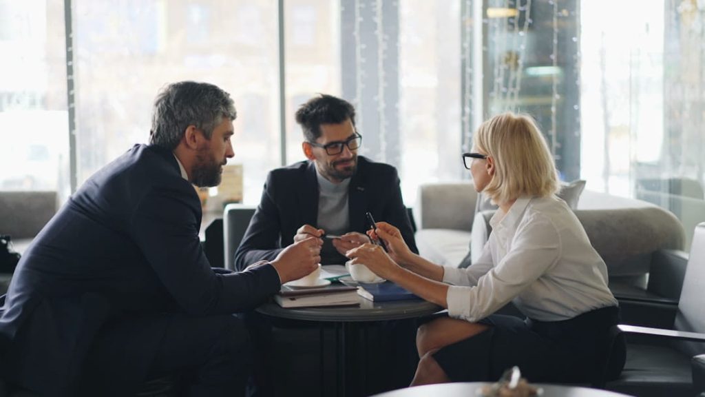 Three business professionals in a comfortable lounge discussing a performance review template over notebooks and coffee.