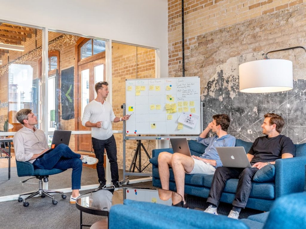 A small team in a casual office running an agile-style meeting around a whiteboard, mapping out steps like those in a performance review template.