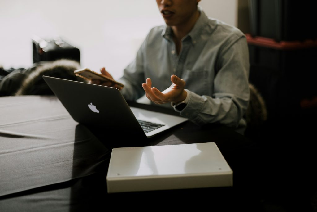 A small business consultant sits at a dark table, engaged with a laptop and a smartphone. They wear a light blue or grey button-up shirt and gesture expressively with both hands, holding a smartphone in the left hand. The dark-colored laptop, likely a MacBook, is open in front of them, with a white rectangular object placed to its right on the table. The background is softly lit on the left, transitioning to darker tones on the right, creating a focused and intimate atmosphere.