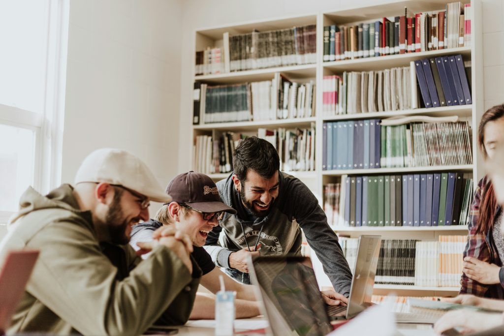 A bright, collaborative study or work environment featuring a group of young adults laughing and engaging with laptops. The central focus is on three men, one wearing a flat cap, another in a baseball cap, and a third in a gray hoodie, all smiling and interacting. A large bookshelf filled with books and binders forms the backdrop, while natural light streams in from a window on the left. The scene conveys a positive and productive atmosphere in an academic or professional setting, much like the energy found at networking events Boston.