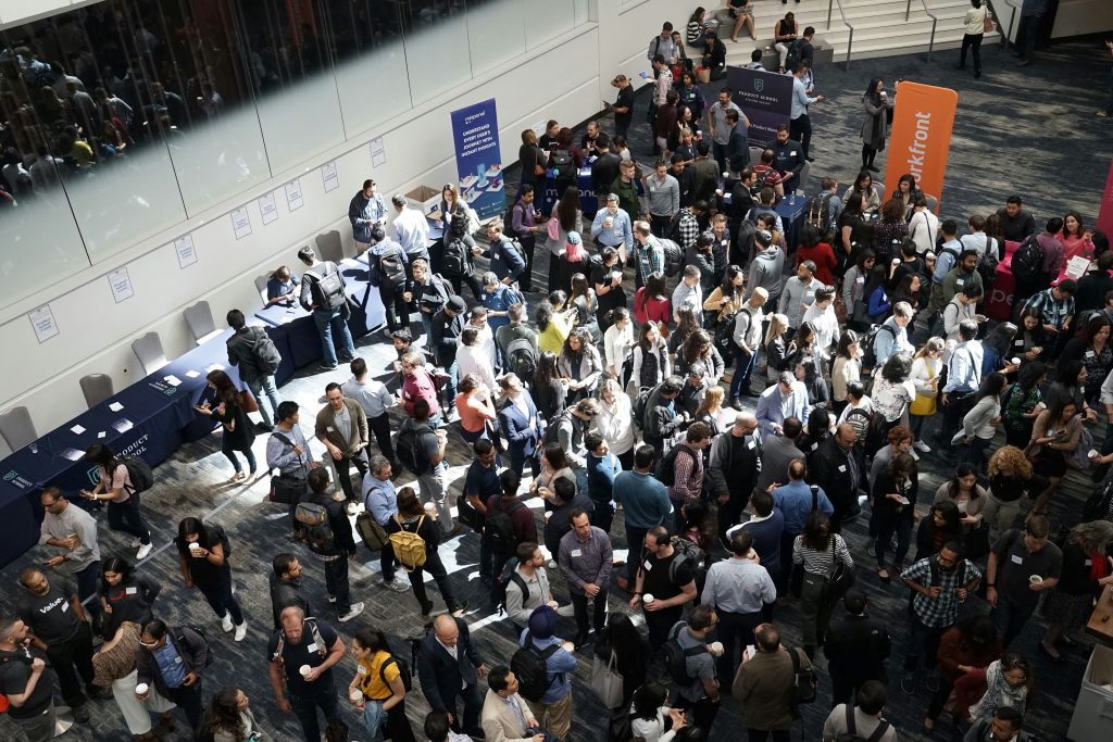 A bustling professional networking events Atlanta or conference, viewed from above, featuring a diverse crowd of attendees mingling and engaging at various sponsor booths and registration tables. The space is modern, with white walls, reflective glass partitions, and a dark gray carpet. Attendees, dressed in business casual attire, are seen holding coffee cups, wearing backpacks, and interacting in small groups, creating a lively and dynamic atmosphere.