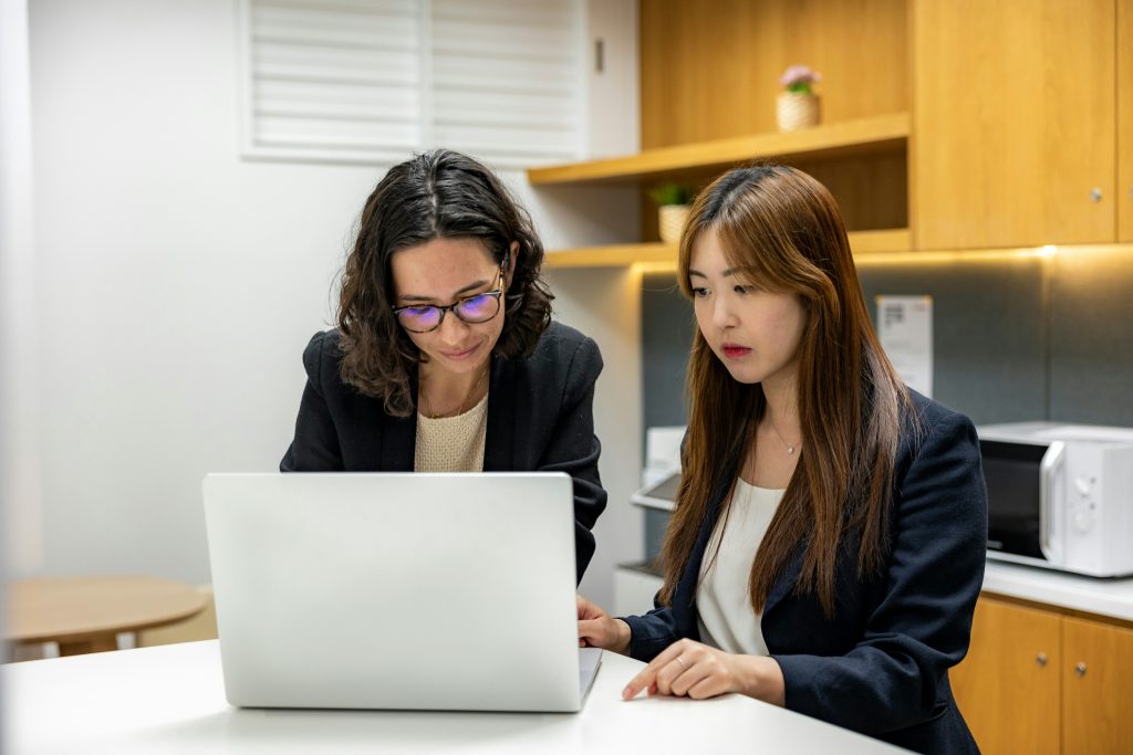 Two women sit at a white table, focused on a silver laptop positioned between them. The woman on the left, with dark curly hair and glasses, wears a light knit top and a dark blazer, while the woman on the right, with long brown hair, wears a white top, a dark blazer, and a silver necklace. The background features a kitchenette with light wooden cabinets, shelves holding small potted plants, and a white microwave on the countertop. The scene is brightly lit, suggesting a professional and collaborative environment, perfect for discussing what are angel matchmaking services.