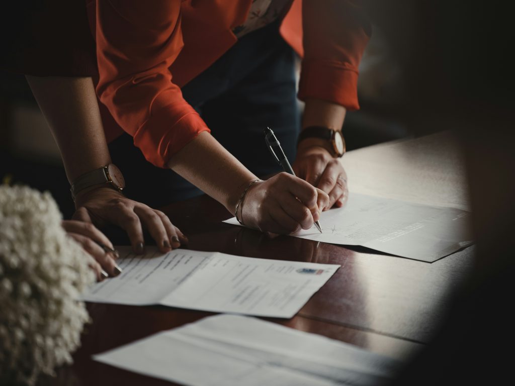 A close-up scene of two people, including a small business lawyer, interacting with documents on a wooden table. One person, wearing a coral-red jacket with rolled-up sleeves, is signing a document with a black and silver pen. Their left hand steadies the paper, while a gold wristwatch and a silver bracelet are visible. Another person's hands, with manicured nails and dark polish, rest on a separate document in the foreground. The setting is dimly lit, with a focus on the hands, pen, and documents, creating a formal and professional atmosphere.