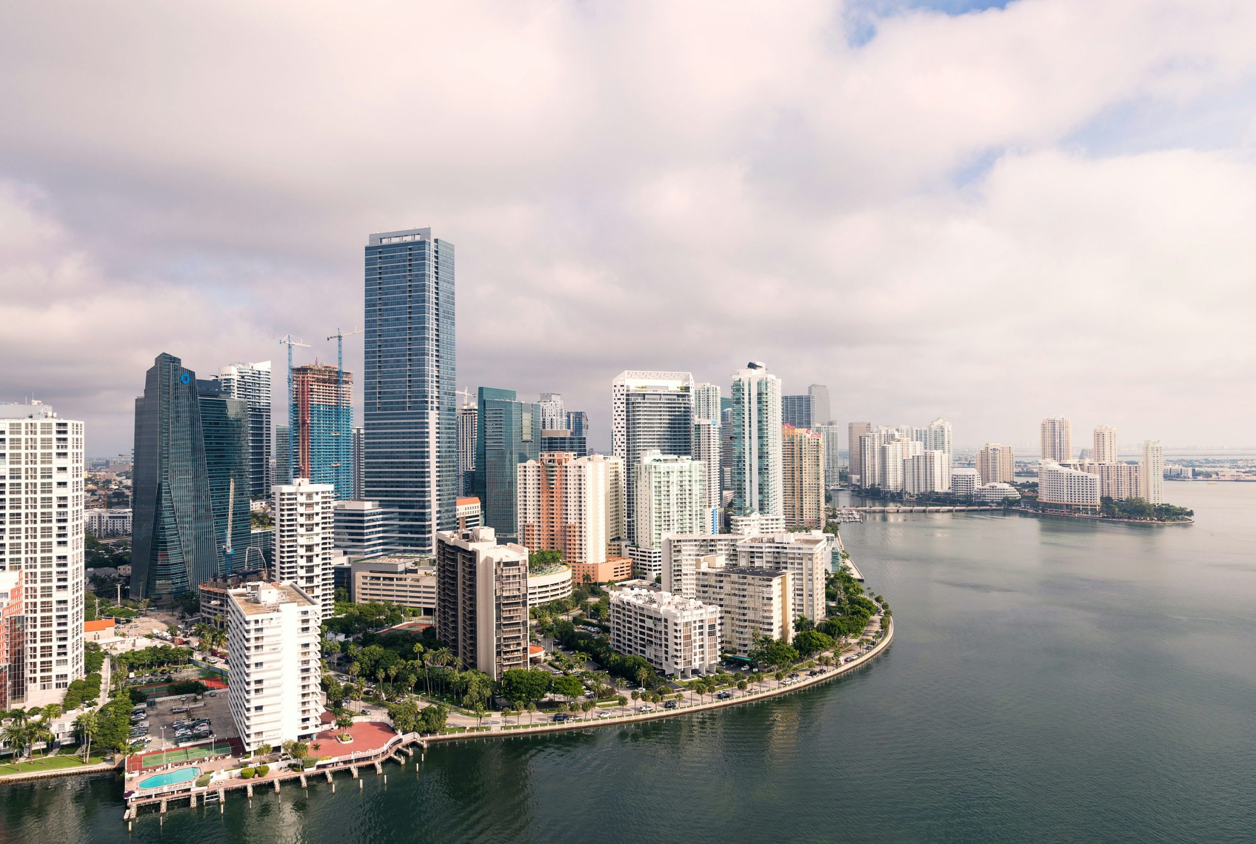 Aerial view of a vibrant city skyline along a curved waterfront under a lightly clouded sky. The city features a mix of modern high-rise buildings, ongoing construction, and lush greenery with palm trees. A calm body of water reflects the skyline, with a curved shoreline lined with buildings and recreational facilities like a swimming pool and tennis courts. Networking events Miami add to the dynamic energy of this bustling environment. A long bridge spans the water in the background, connecting landmasses. The scene evokes a bustling coastal city, likely in a subtropical climate.