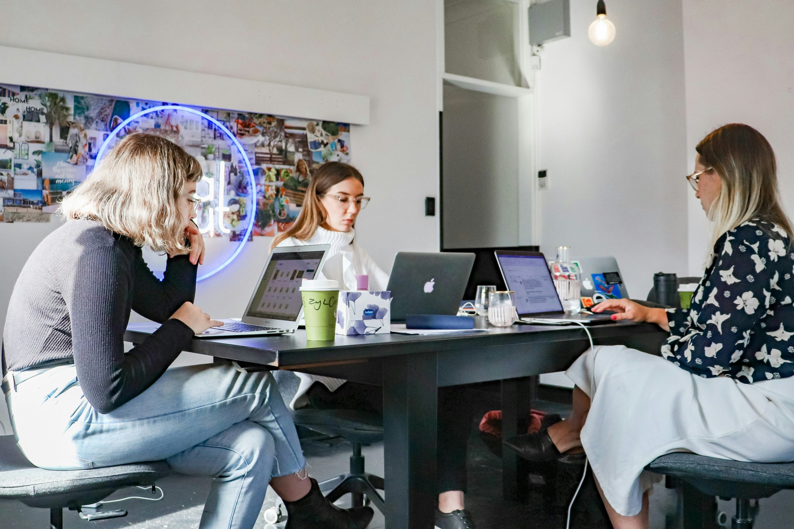 Three women from one of the cheap small business marketing agencies working on laptops at a dark rectangular table in a brightly lit, modern office space. The table is filled with laptops, drinks, and other items, including a light green paper cup and a tissue box. Behind them, a wall collage of photos and text is partially covered by a circular blue neon sign. The women are seated on dark office chairs, focused on their work, with a creative and collaborative atmosphere evident in the setting.