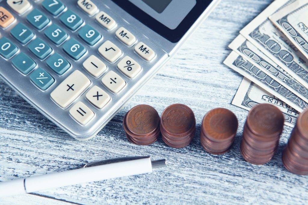 A financial-themed arrangement on a light wooden surface featuring a silver calculator with visible numeric and function keys, including memory and arithmetic operations, perhaps to calculate how much do brokers charge to sell a business. To the right, several U.S. one hundred dollar bills are fanned out, partially overlapping. Below the calculator, five neatly stacked piles of copper-colored coins are aligned horizontally, with "MEXICO" visible on one coin. A white pen with a metallic clip lies horizontally in the bottom left corner. The scene conveys a focus on budgeting, accounting, or financial calculations.