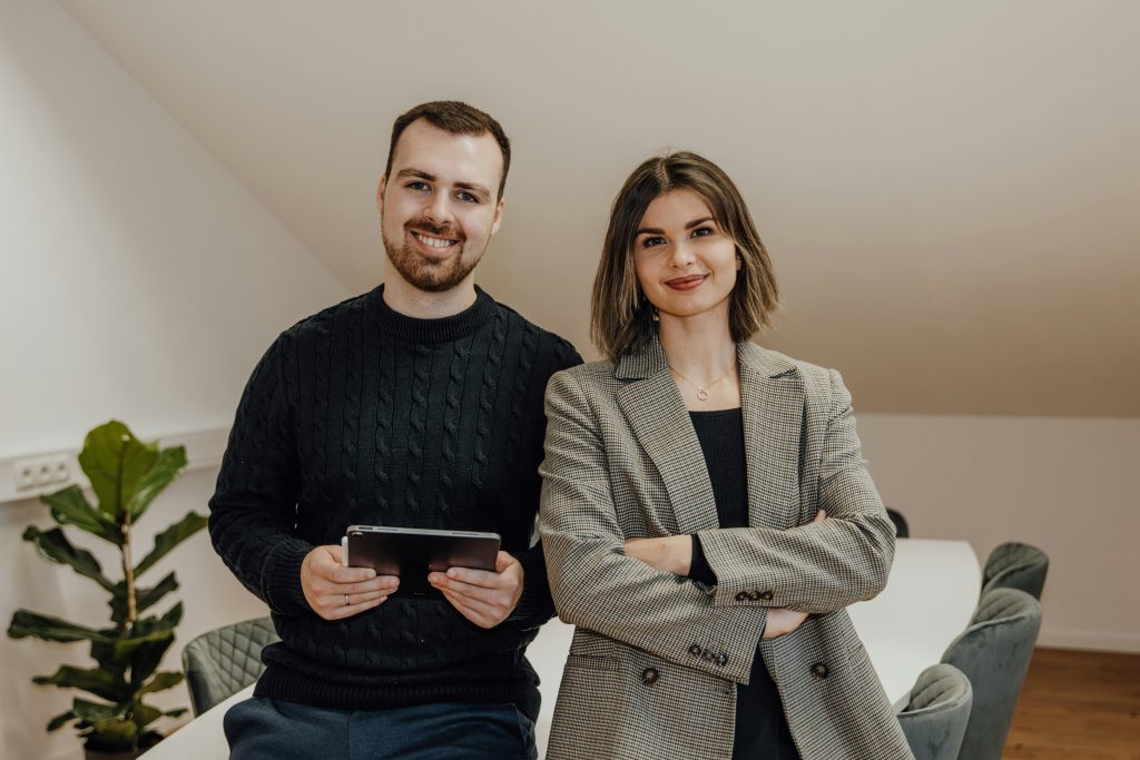 Two smiling professionals, a man and a woman, standing in a bright, modern office setting. The man, on the left, has short brown hair, a beard, and is wearing a dark cable-knit sweater while holding a tablet. The woman, on the right, has a bob haircut with highlights, is dressed in a houndstooth-patterned blazer over a dark top, and has her arms crossed. The background features light-colored walls, a sloped ceiling, a green potted plant, and parts of a table and chairs, creating a professional and welcoming atmosphere, perfect for discussing cheap small business marketing agencies.