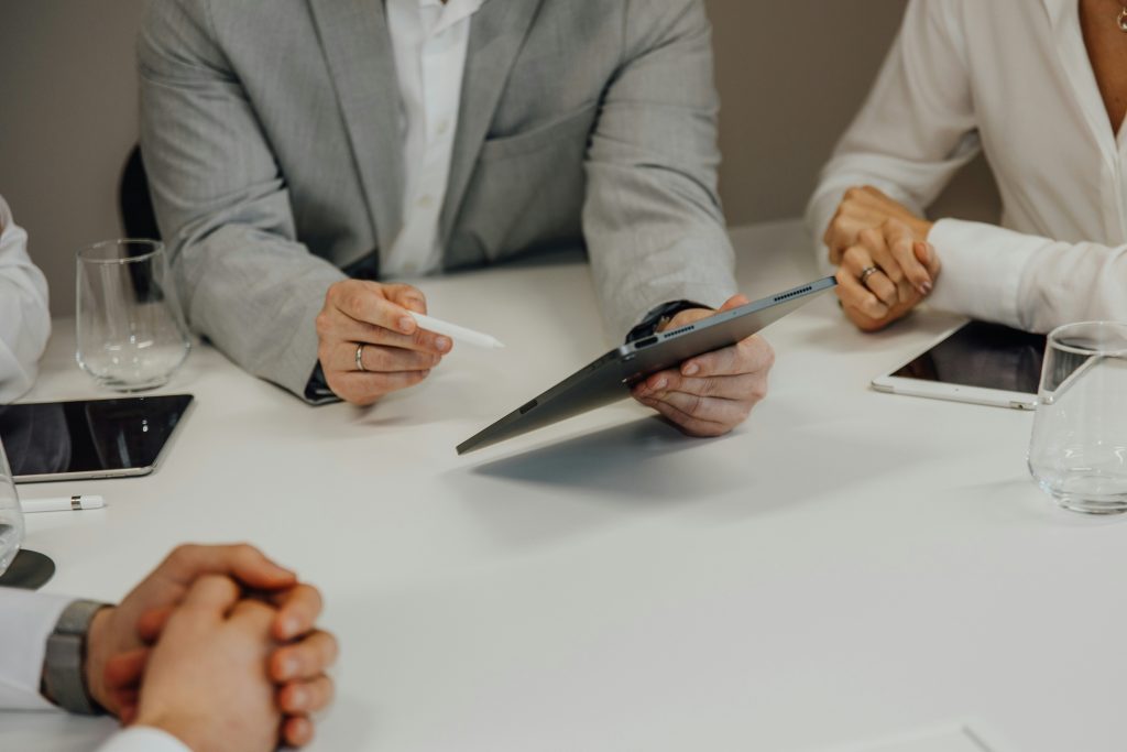 Close-up view of a business meeting with several individuals seated around a white table. The central figure, wearing a light grey suit jacket, holds a dark grey tablet and a white stylus. To the right, another person in a white shirt has clasped hands near a silver tablet and a clear glass tumbler. On the left, part of another person is visible with a similar glass and a dark tablet. The table features multiple tablets, styluses, and glasses, emphasizing a collaborative and professional setting, ideal for cheap small business marketing agencies to strategize and connect.