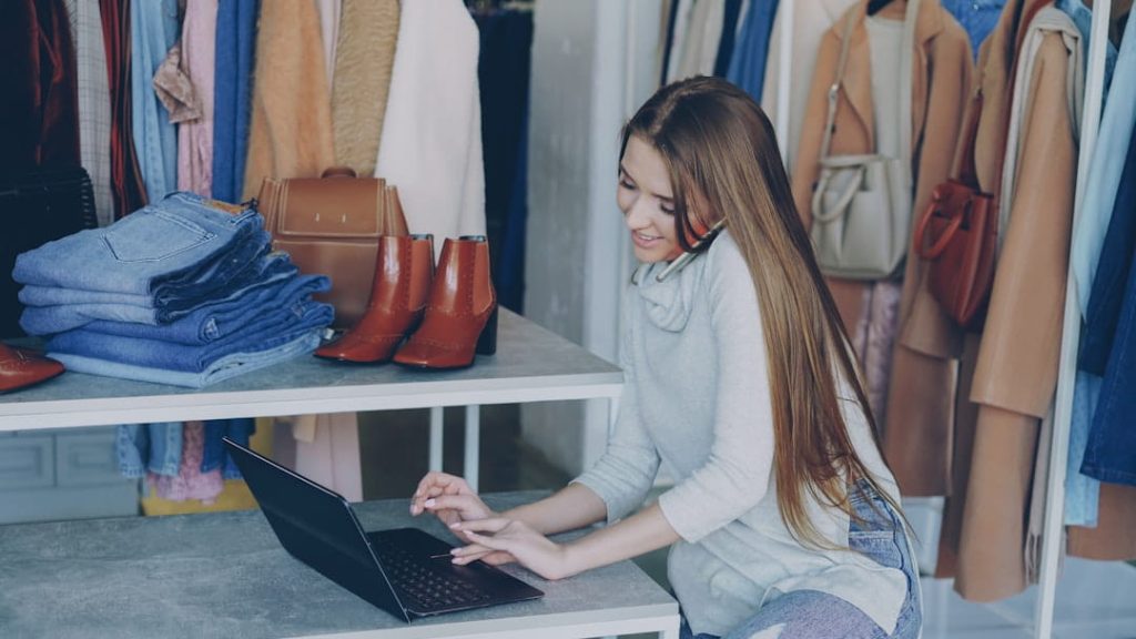 Woman in a clothing boutique typing on a laptop while chatting on the phone, managing her store and her Shopify SEO checklist.
