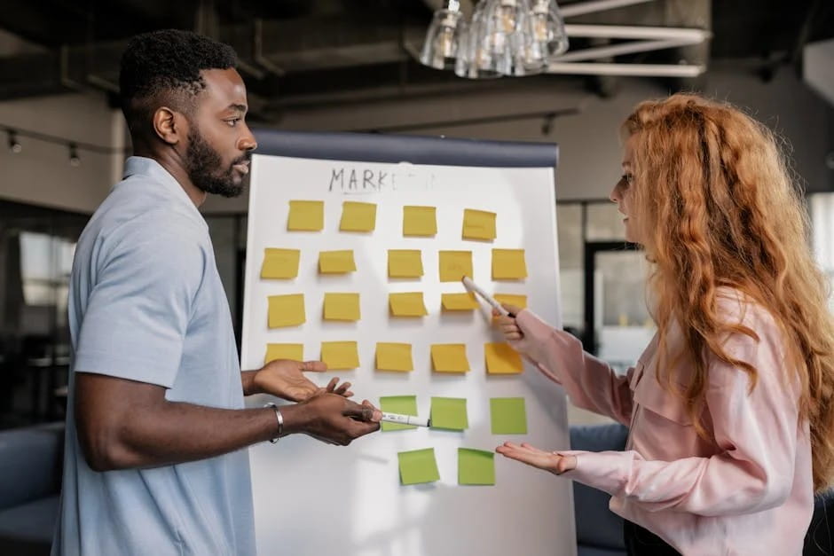 Two colleagues, a man and a woman, collaborate on a marketing brainstorming session using a whiteboard filled with yellow and green sticky notes in a modern office during Small Business Month.