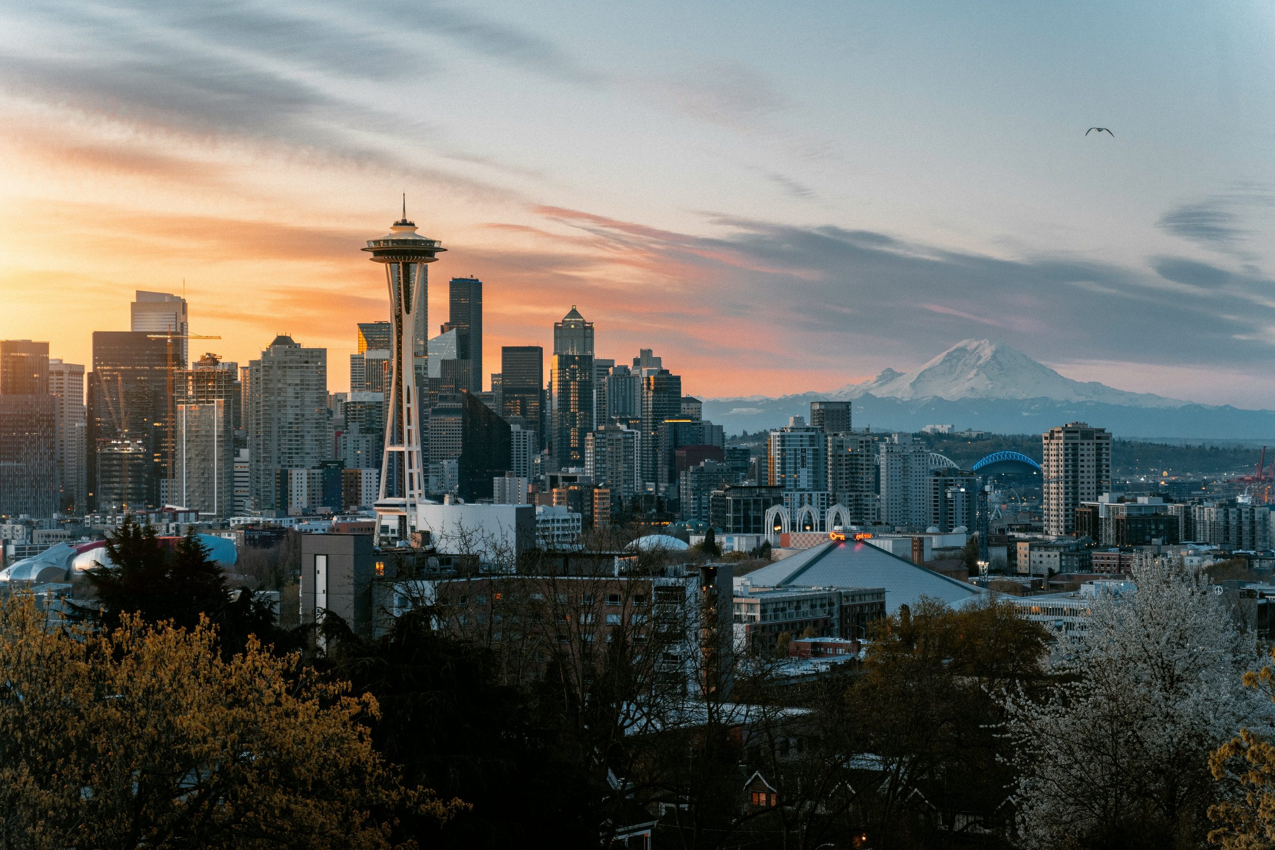 Panoramic view of the Seattle skyline at sunrise or sunset, featuring the iconic Space Needle surrounded by high-rise buildings. The cityscape offers networking events Seattle framed by lush green trees in the foreground and the majestic, snow-capped Mount Rainier in the background under a gradient sky transitioning from warm orange to cool blue hues.