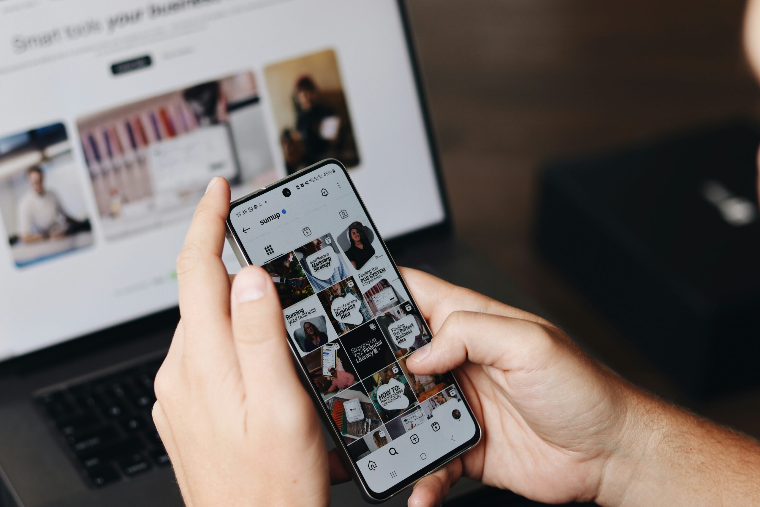 Close-up of hands holding a smartphone displaying an Instagram feed for the account 'sumup,' featuring posts about small business strategies, financial literacy, and marketing. The phone screen shows a grid of text-heavy posts with topics like 'Small Business Marketing Strategy' and 'Finding the Perfect Business Idea.' A laptop is visible in the blurred background, partially showing the text 'Smart tools your business,' reinforcing the theme of business tools and resources for ads for small business.
