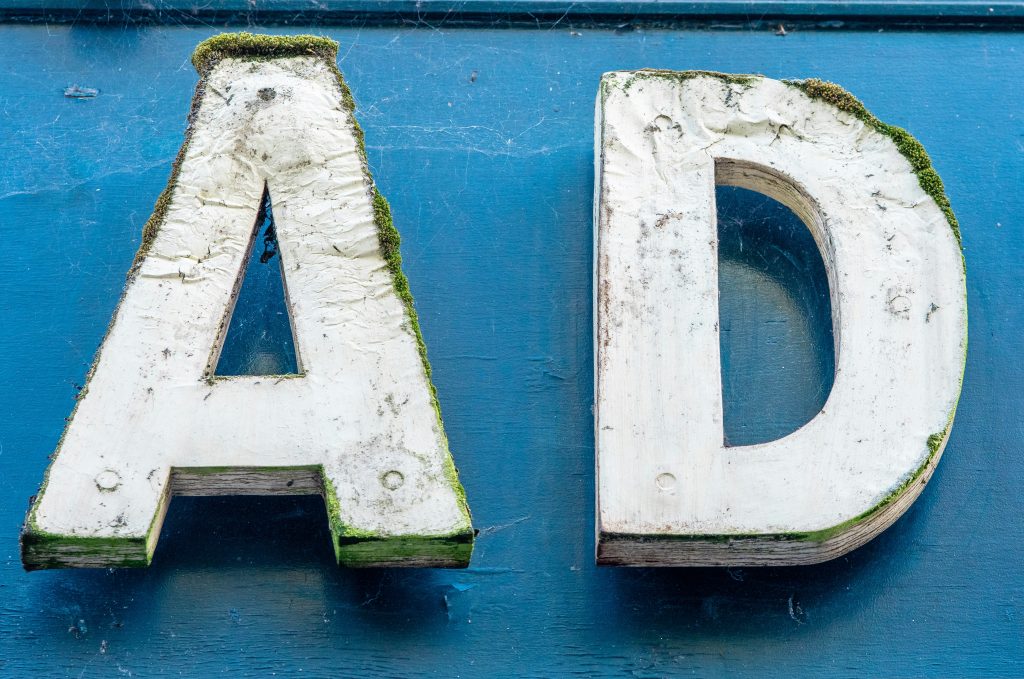 Close-up of two large, weathered wooden letters, 'A' and 'D', painted white with visible cracks, peeling paint, and green moss growth along the edges. The letters are set against a solid blue background with faint textures and subtle spiderwebs, evoking a rustic and aged aesthetic, reminiscent of ads for small business charm.