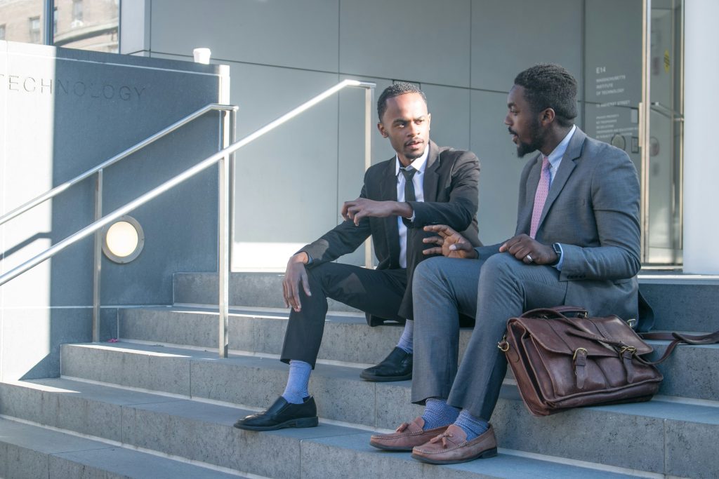 Two men in business attire seated on concrete steps outside a building, with a brown leather bag, and coffee cup, discussing conferences in Las Vegas this week.