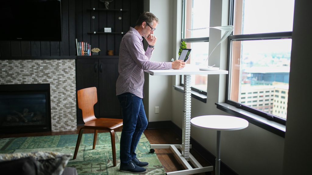 A man holds a tablet, standing thoughtfully at a white desk in a modern living room or office as he considers virtual meeting etiquette. The room has a dark wood-paneled wall with a TV over a fireplace, built-in shelves, and large windows with a city view. The minimalist desk holds a lamp and plant. A patterned rug and wooden chair add warmth to the functional, contemporary space.