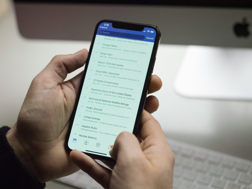 A person holds a black smartphone vertically with both hands, displaying a news feed or search results for networking events in DC on the screen. The bottom navigation bar features five icons, with a red notification badge showing "9" on the bell icon. The background is slightly blurred, revealing part of an Apple monitor, a white desk, and a keyboard, suggesting an office or personal workspace. The focus is on the phone and its content, highlighting mobile news consumption.